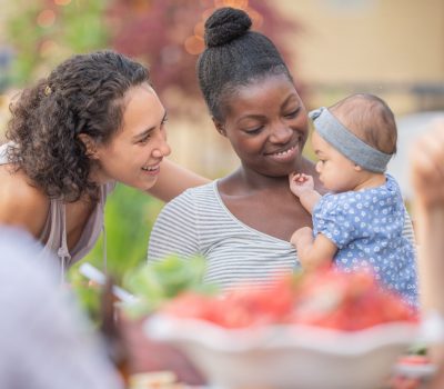A young African American mom holds her adorable young daughter in her lap at an outdoor dining table. The girl is smiling and reaching toward mom's friend, who's smiling at her.